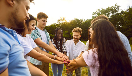 Multiracial men and women outdoors in summer. Group of cheerful young friends stacking their hands together while having fun in park. Human relationship, social, community and team building concept.の写真素材