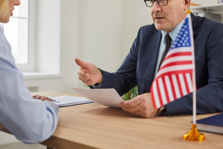 USA consulate worker sitting at office table with American flag and explaining woman customer...