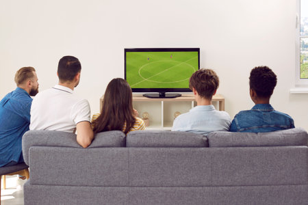 Multinational friends - fans gathered together, sitting on sofa in large room to watch a football...