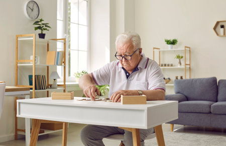Chubby old senior retired man in glasses sitting at desk, playing games, solving puzzles and keeping his brain young. Prevention of dementia and Alzheimers disease conceptの写真素材