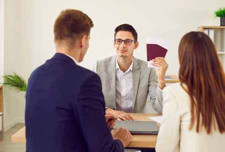 Young couple meeting with a travel agent or visa manager. Male agency worker sitting at his office desk with a young man and woman, looking at them and holding their tickets and passportsの写真素材