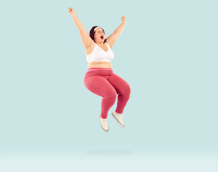 Full length photo of a funny overjoyed young fat woman wearing sportswear jumping with excited expression, open mouth and hands up isolated on studio blue background. Workout, gym, fitness concept.の写真素材