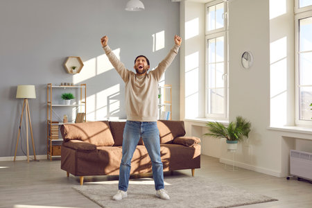 Free, happy, excited man having fun at home. Funny, joyful, crazy adult man having a day off, enjoying freedom, standing alone in the living room, raising his hands up, and screamingの写真素材