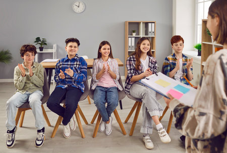 Group of smiling school children clapping their hands while sitting in row in classroom. Schoolgirl standing in front of her classmates acting as teacher. School education, discussionの写真素材