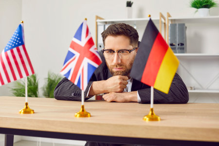 Thoughtful young man sitting at desk in office looking at USA, Great Britain and Germany flags. Businessman choosing foreign language or country to work or study in. Education or emigrationの写真素材