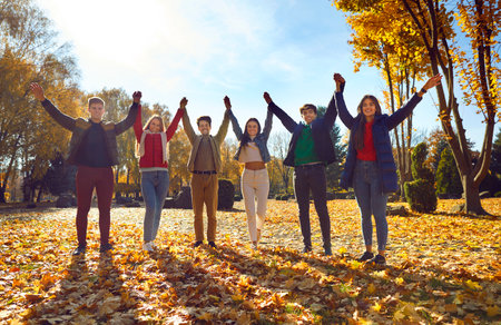 Happy friends standing together raising their hands in park. Group of young people wearing warm...