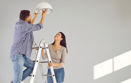 Happy loving couple changing light bulb in new apartment. Family moving in new house, doing DIY renovations at home. Cheerful husband and wife standing on ladderの写真素材