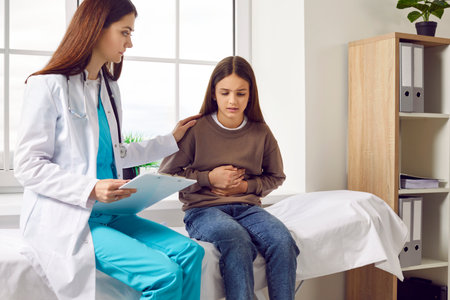 Teenage girl sitting on the couch in the doctors office and pointing to her stomach to paediatrician...