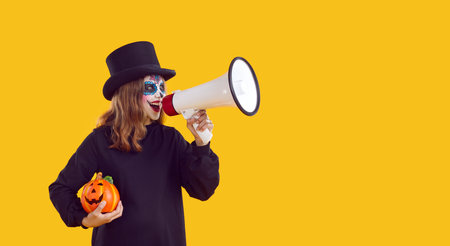 Happy child girl in spooky Halloween costume standing isolated on yellow color copyspace background holding pumpkin and megaphone, making announcement, advertising festive event, party or holiday saleの写真素材