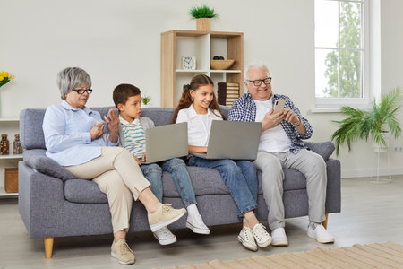 Happy grandparents with grandchildren girl and boy sitting on sofa using modern laptops and smartphones together. Smiling seniors resting on the couch at home with kids enjoying weekend.の写真素材