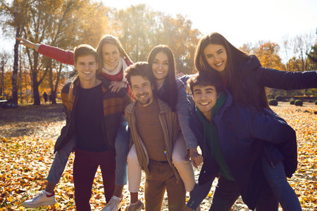 Group of joyful young friends having fun in a sunny autumn park. Several happy smiling young people piggybacking their girlfriends while walking in bright sun light on a nice day in autumnの写真素材