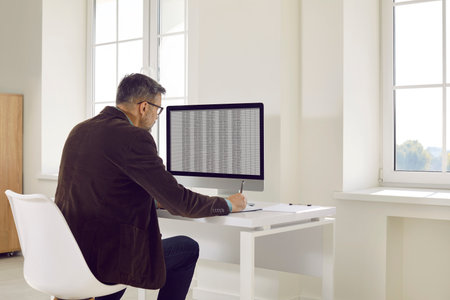 Mature business accountant or auditor working in office. Back view of senior man sitting at desk, looking at computer screen, using digital financial spreadsheets and talking notes with penの写真素材