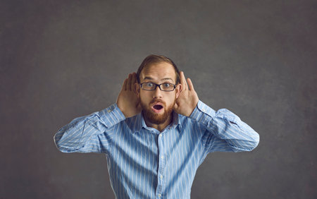 Funny curious young man standing in on grey studio background looking at camera with open mouth and...