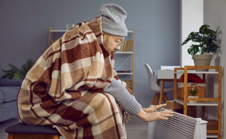 Senior man freezing and using an electric heater in winter. Old man wrapped in a plaid blanket sitting in a chilly room at home, feeling cold and warming up his hands by his modern electric heaterの写真素材