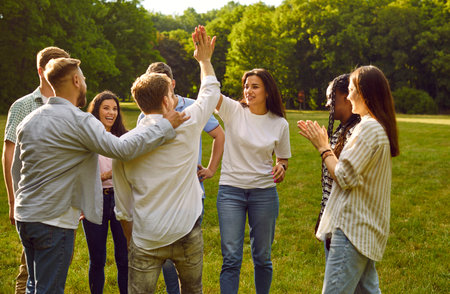 Team of happy, joyful diverse friends meet in the park on a good summer day. Happy man and woman give each other a high five while other young people are clapping hands. Teamwork conceptの写真素材