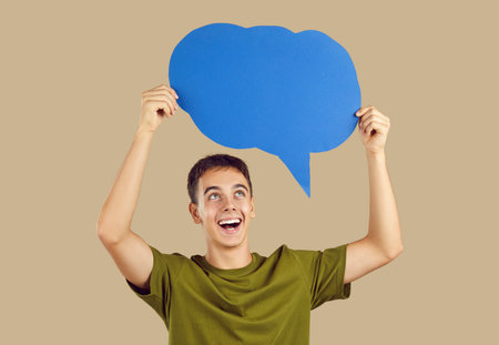 Happy excited young boy wearing green t-shirt holding blank speech bubble for thoughts isolated on...