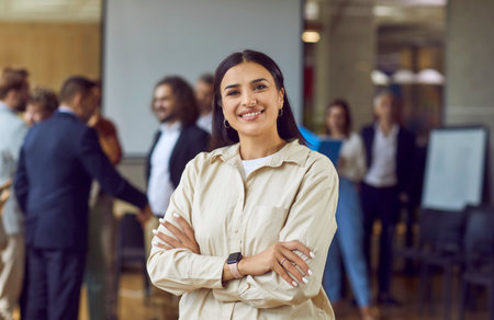 Close up portrait of a happy smiling confident young business woman leader proudly looking at the camera with crossed arms with a team of company employees talking in background in modern office.の写真素材