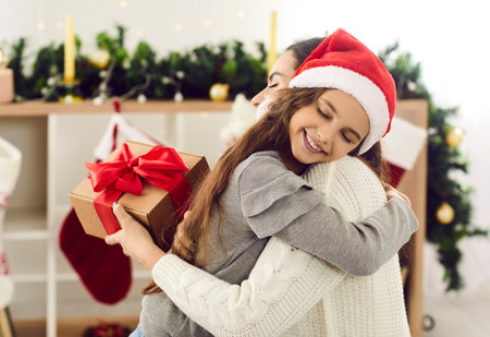 Parents and children exchange gifts. Cute little girl tenderly hugs her mother wishing her Merry Christmas and Happy New Year. Side view of girl in Santa hat with closed eyes in arms of her mother.の写真素材