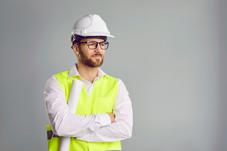 Man builder in hardhat standing with blueprints in his hands. Construction company employee wearing safety vest and helmet posing with his arm folded against gray wall. Engineering construction workerの写真素材