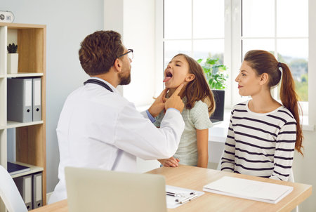 Male doctor otolaryngologist examining teenage girl patients throat in medical office during checkup. Doctor pediatrician exam the oral cavity of sick child sitting with mother in examination room.の写真素材