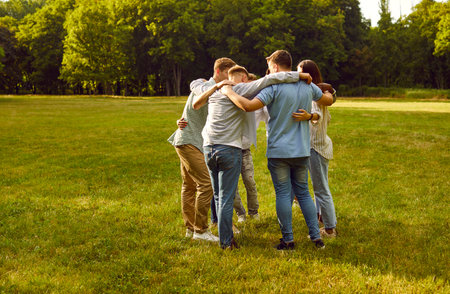 Group of people standing in circle in middle of green park and hugging each other. Young men and women are chatting in circle during meeting outdoors. Close friendship and bonding concept.の写真素材