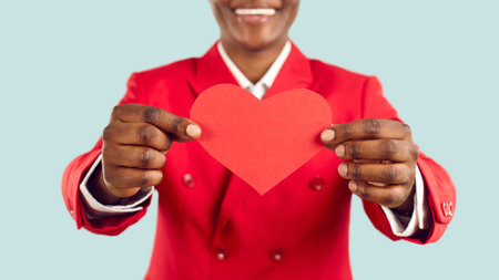 Happy handsome young African American man in red suit holding red heart shaped paper card in hands. Close up. Cropped shot. Concept of St Valentines Day celebrationの写真素材