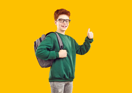Happy funny red-haired schoolboy with glasses, dressed in green sweater and gray jeans with backpack on his back shows thumbs-up - cool, photo on isolated yellow background. School education concept.の写真素材