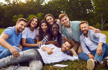 Portrait of cheerful students posing while sitting in park on grass in summer. Funny smiling multiracial men and women spending time together in nature after studying. Education and friendship conceptの写真素材