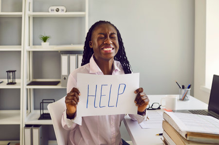 African american stressed upset overworked woman sitting at the desk at her workplace in office with a pile of folders. Tired business girl holding a sign with the inscription word help.の写真素材