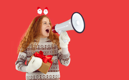Happy curly friendly redhead girl holding Christmas gift present in hands shouting loudspeaker on red background. Christmas, New Year banner for marketing, sales, announcement and notification.の写真素材