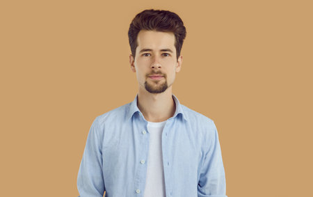 Portrait of smiling young man with neatly shaved beard and mustache, dressed in white T-shirt and blue shirt, confidently looking straight into the camera. Studio photo on isolated beige background.の写真素材