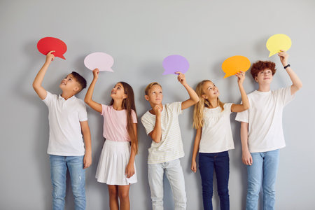 Children sharing opinions using speech bubbles and thinking clouds. Group of kids standing by grey studio wall looking at colourful mockup cards in their hands with happy or pensive face expressionsの写真素材