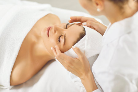 Close up portrait of a beautiful young woman lying with closed eyes and having face or head massage in spa. Woman relax enjoying health therapy treatment. Wellness and beauty day concept.の写真素材