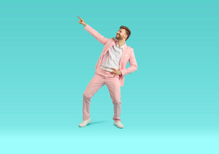 Happy young man in pastel pink trendy suit dancing. Full body length portrait of stylish excited bearded man pointing finger to copy space over isolated light blue studio backgroundの写真素材