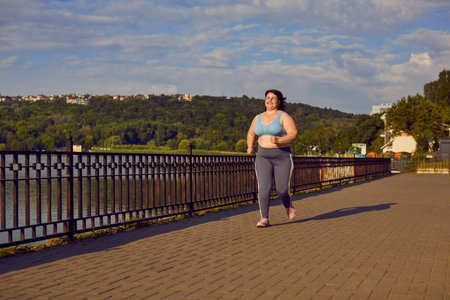 Overweight woman having a jogging workout in the city. Happy fat chubby girl in sportswear running on a bridge over the river, with a beautiful landscape in the background. Sport, fitness conceptの写真素材