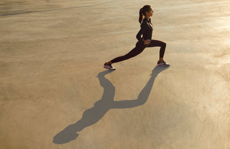 Woman doing outdoor morning workout on city street. Fit young girl in black sportswear doing forward lunges on gray sunlit urban concrete floor. Sport, exercise, stretching conceptの写真素材