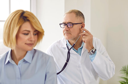 Mature smiling female patient in clinic for a medical checkup. Senior physician doctor with stethoscope examining womans lungs, breathing and heartbeat at the hospital in exam room.の写真素材