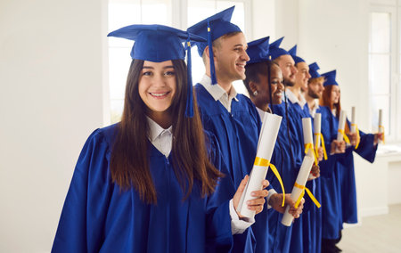 Portrait of young smiling joyful girl student in university graduate gown and diploma in hands. Happy graduated woman standing with classmates indoors. Graduation celebration and education concept.の写真素材