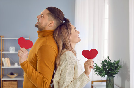 Portrait of a young happy smiling man and woman standing back to back and holding red paper hearts in their hands at home. Happy couple in love. Care, tenderness and Valentines day concept.の写真素材