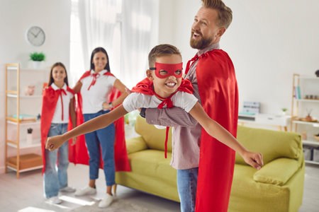 Happy smiling family of four in red super hero costumes playing game imagine flying at home. Young man parent lifting a boy child in air playing plane enjoying spending time together.の写真素材