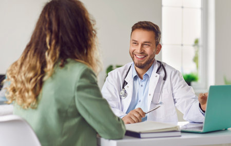 Cheerful friendly male doctor at medical office in hospital, showing examination results on laptop monitor screen and having consultation with young woman patient sitting in clinic.の写真素材