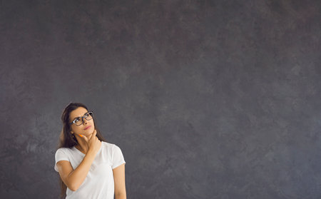 Pensive millennial woman on black studio background look at blank empty copy space thinking making...
