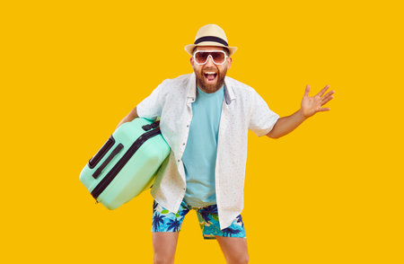 Happy male tourist with suitcase. Joyful bearded man in summer clothes, cool sunglasses and fedora sunhat holding his bag and standing isolated on yellow color background. Travel, holiday conceptの写真素材