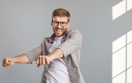 Happy dancing man. Portrait of a cheerful, joyful, smiling young guy in a casual shirt and glasses, with a short beard dancing on a grey wall background. Party, having fun conceptsの写真素材