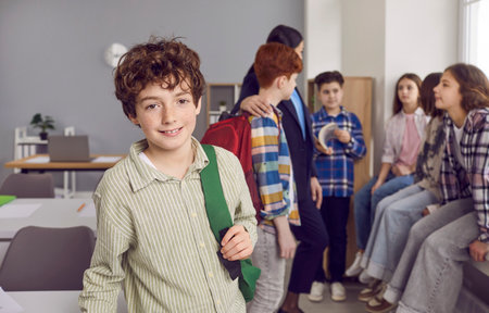 Portrait of smiling cute boy standing with backpack in classroom. Happy schoolboy wearing casual clothes standing on background of his classmates and teacher and looking at cameraの写真素材