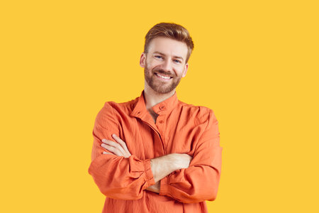 Portrait of confident smiling young bearded handsome man standing with crossed arms wearing casual bright orange summer shirt isolated on studio yellow background. Smart guy looking at the camera.の写真素材
