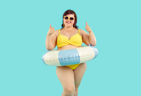 Cheerful chubby woman with swimming ring showing thumbs up on pastel light blue background. Portrait of happy overweight Caucasian woman wearing swimsuit and sunglasses recommending something. Banner.の写真素材