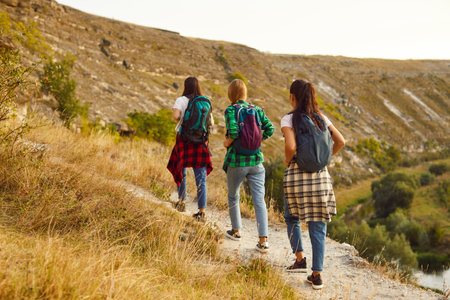 Back view of a group of young girls tourists with backpacks hiking in nature walking in a row....