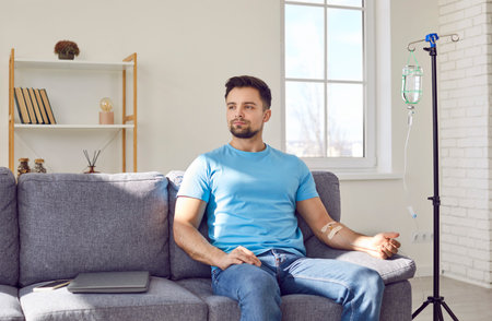 Male patient undergoing vitamin therapy. Handsome young man sitting on the sofa at home and getting vitamin supply through an intravenous medical IV line infusionの写真素材