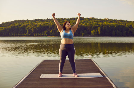 Young happy smiling funny fat woman in sportswear do winners gesture holding hands up in fists celebrating success in workout standing outdoors in the park on pier. Sport and fitness concept.の写真素材